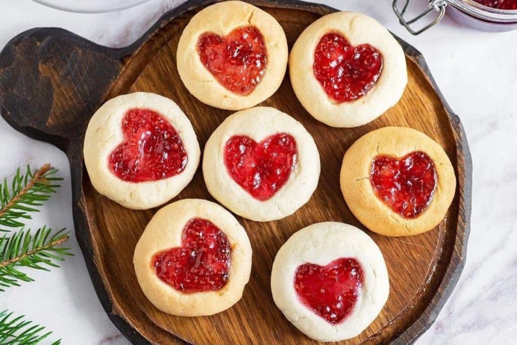 Heart thumbprint cookies filled with raspberry jam served on a round wooden board. These are one of our delcious cookies for Valentine's Day.